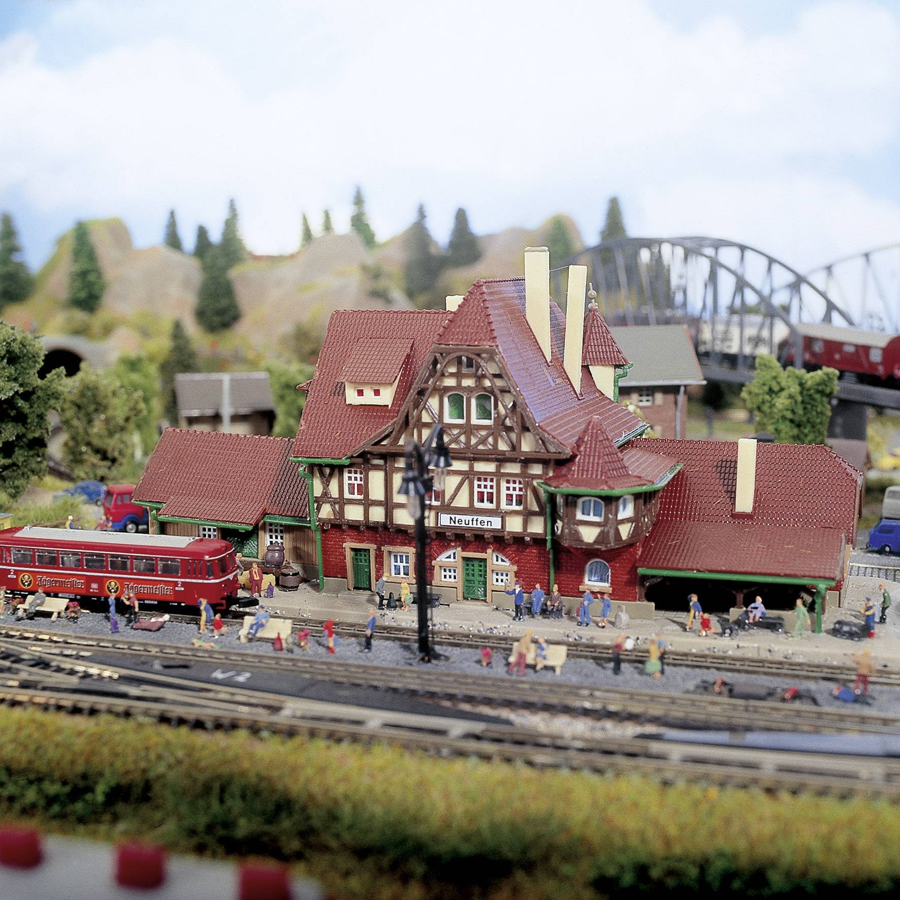 Eine Miniatureisenbahnanlage mit einem Bahnhof, umgeben von Bäumen und Bergen. Ein roter Personenwagen und viele Figuren sind zu sehen.
