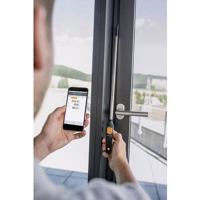 Eine Person misst die Feuchtigkeit eines Fensters mit einem Gerät, das die Werte an ein Smartphone überträgt. Hintergrund: Landschaft.