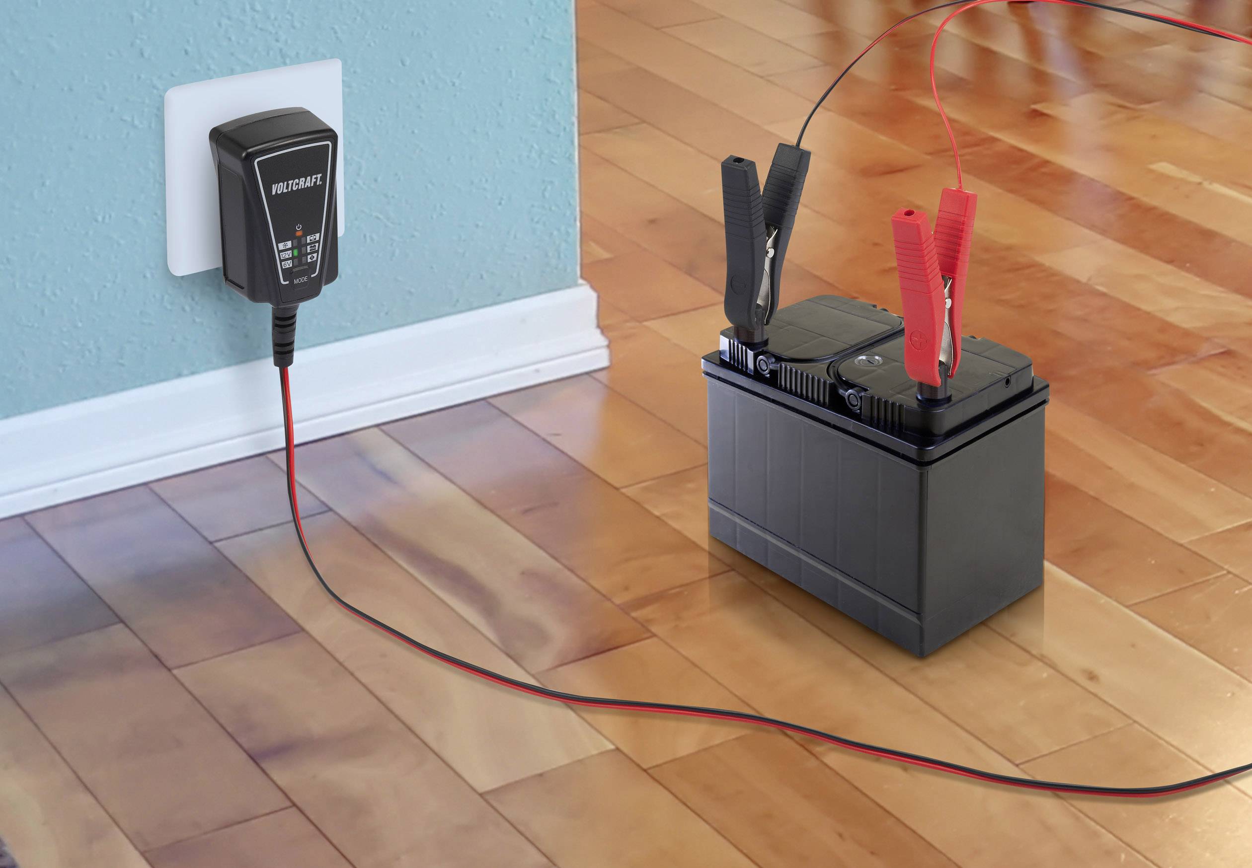 Ein Batterieladegerät ist an einer Wandsteckdose angeschlossen und lädt eine Autobatterie über rote und schwarze Klemmen auf einem Holzboden.