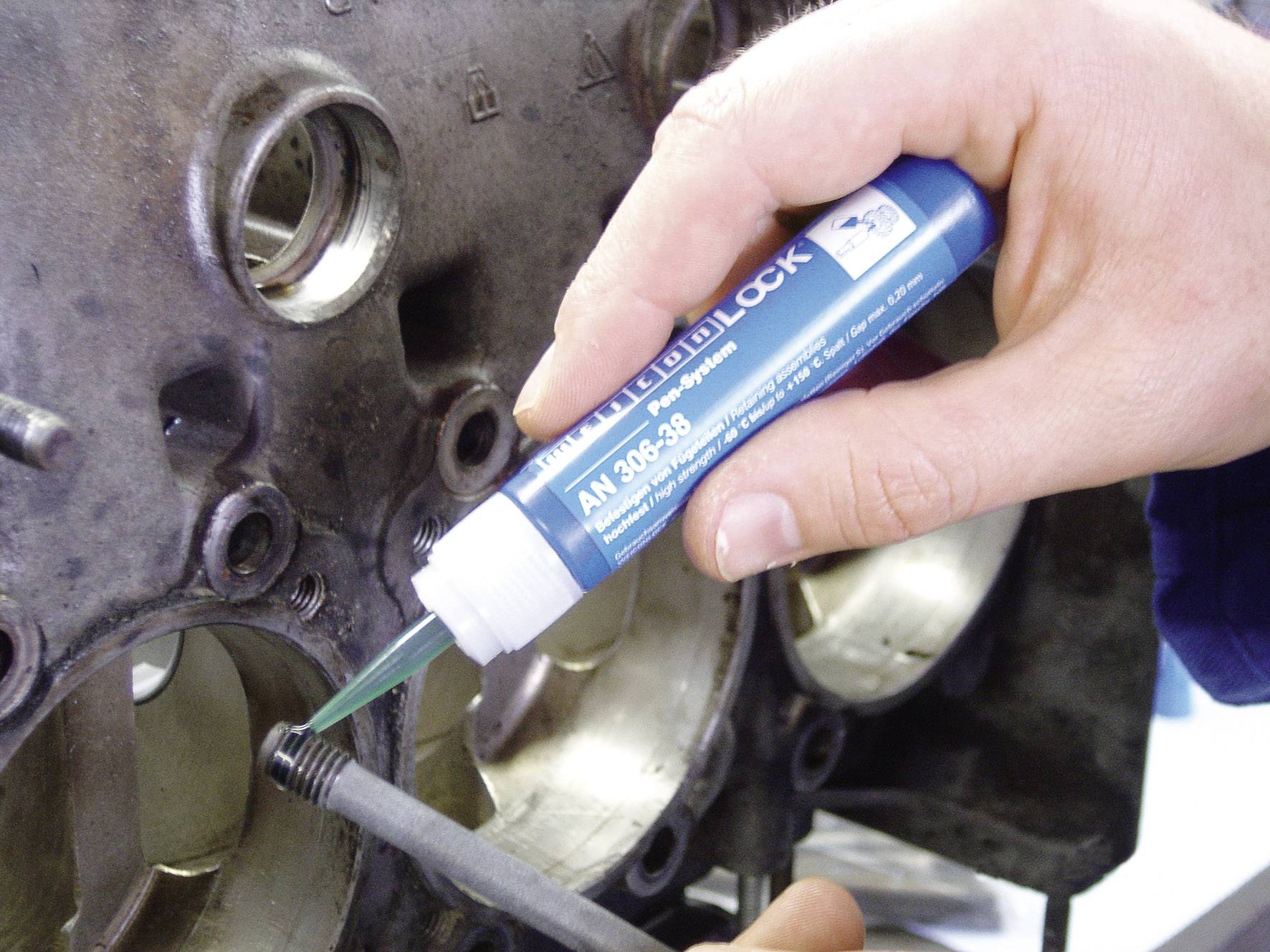 Eine Hand trägt blaue Flüssigkeit aus einer Tube auf eine Schraube in einem Metallteil auf, wahrscheinlich zur Befestigung.