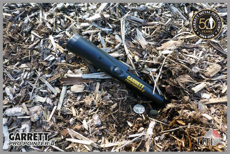 Ein schwarzer Metalldetektor der Marke 'Garrett Pro-Pointer II' liegt auf einem Boden aus Holzschnitzeln. Daneben liegt eine Münze.