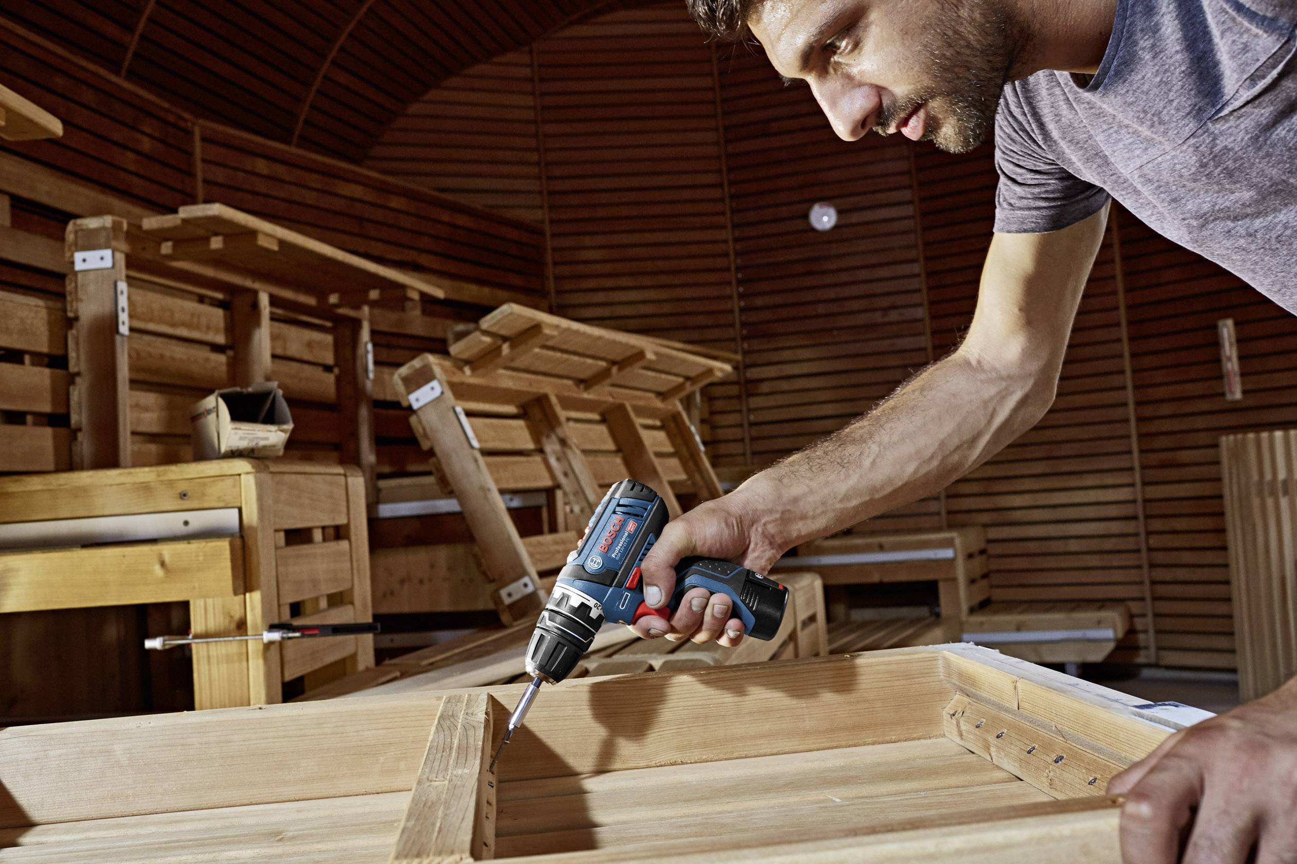 Ein Mann nutzt einen Akkuschrauber, um ein Holzprojekt in einer Werkstatt zu bearbeiten. Im Hintergrund sind Holzmöbel zu sehen.