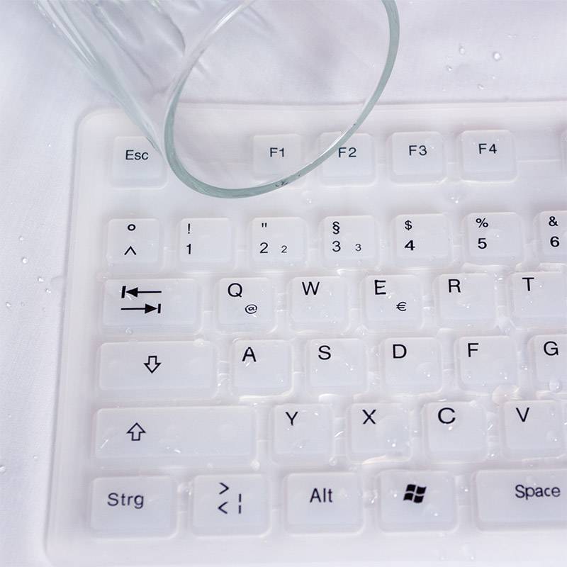 Eine Tastatur auf einem Tisch mit Wassertropfen und einem umgestürzten Glas darauf, zeigt Feuchtigkeit und eine mögliche Verschüttung.