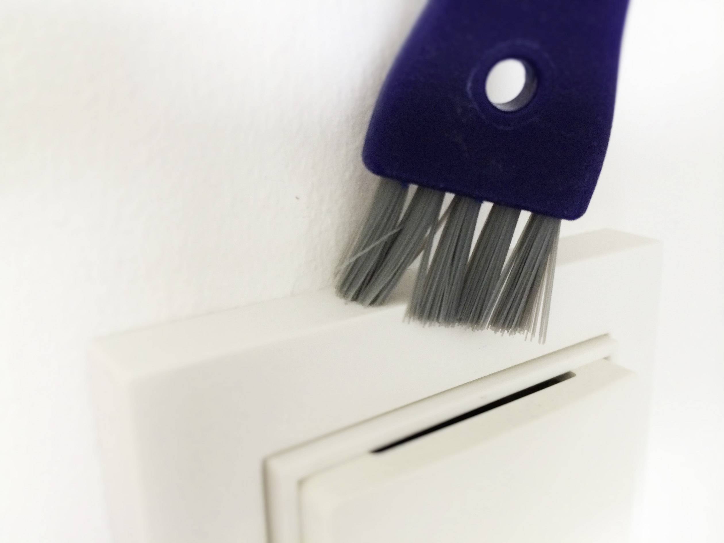 Eine blaue Bürste wird verwendet, um Staub von einem weißen Lichtschalter an einer weißen Wand zu entfernen.