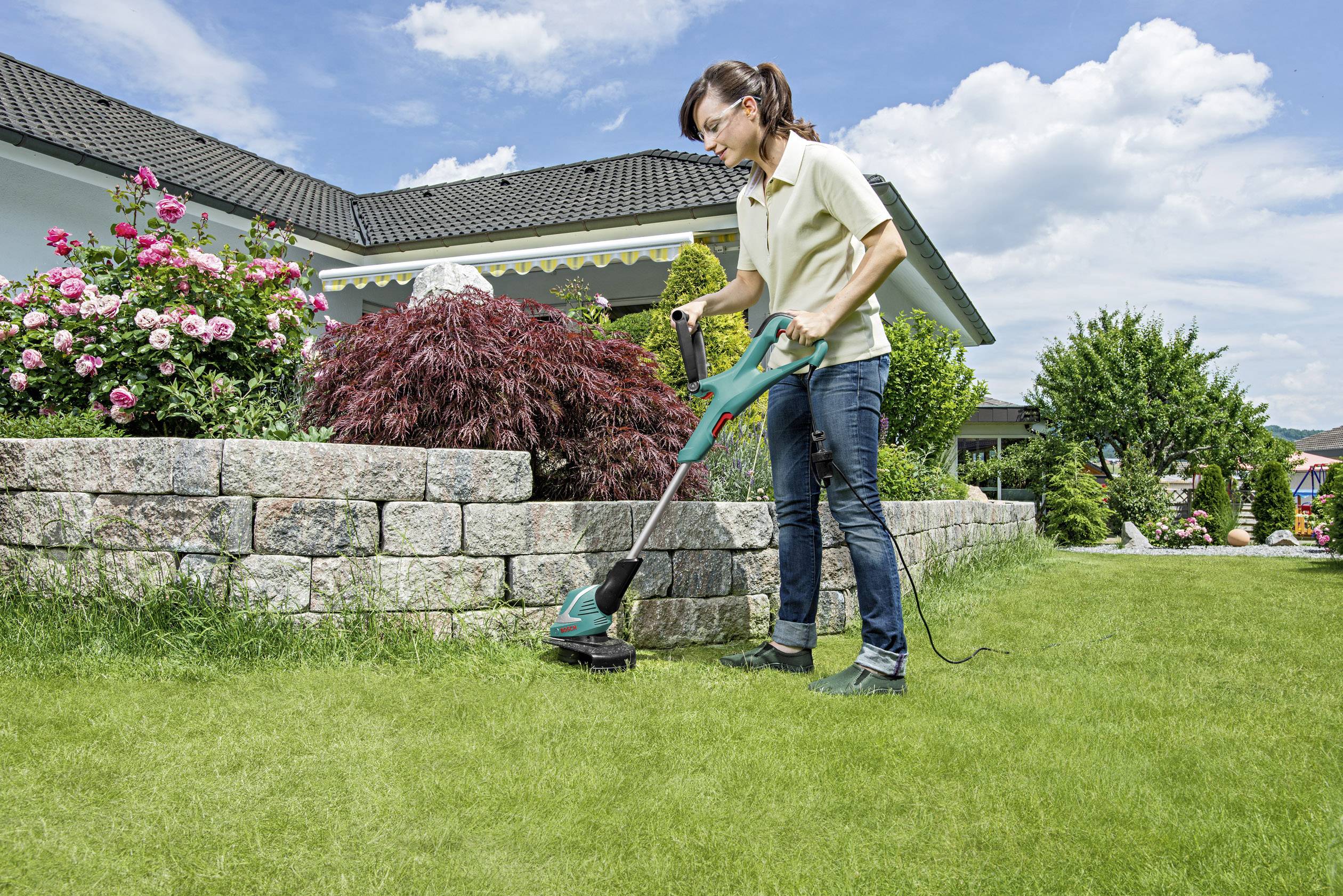 Eine Person mäht den Rasen mit einem elektrischen Trimmer in einem gepflegten Garten vor einem Haus mit Blumenbeeten und Hecken.