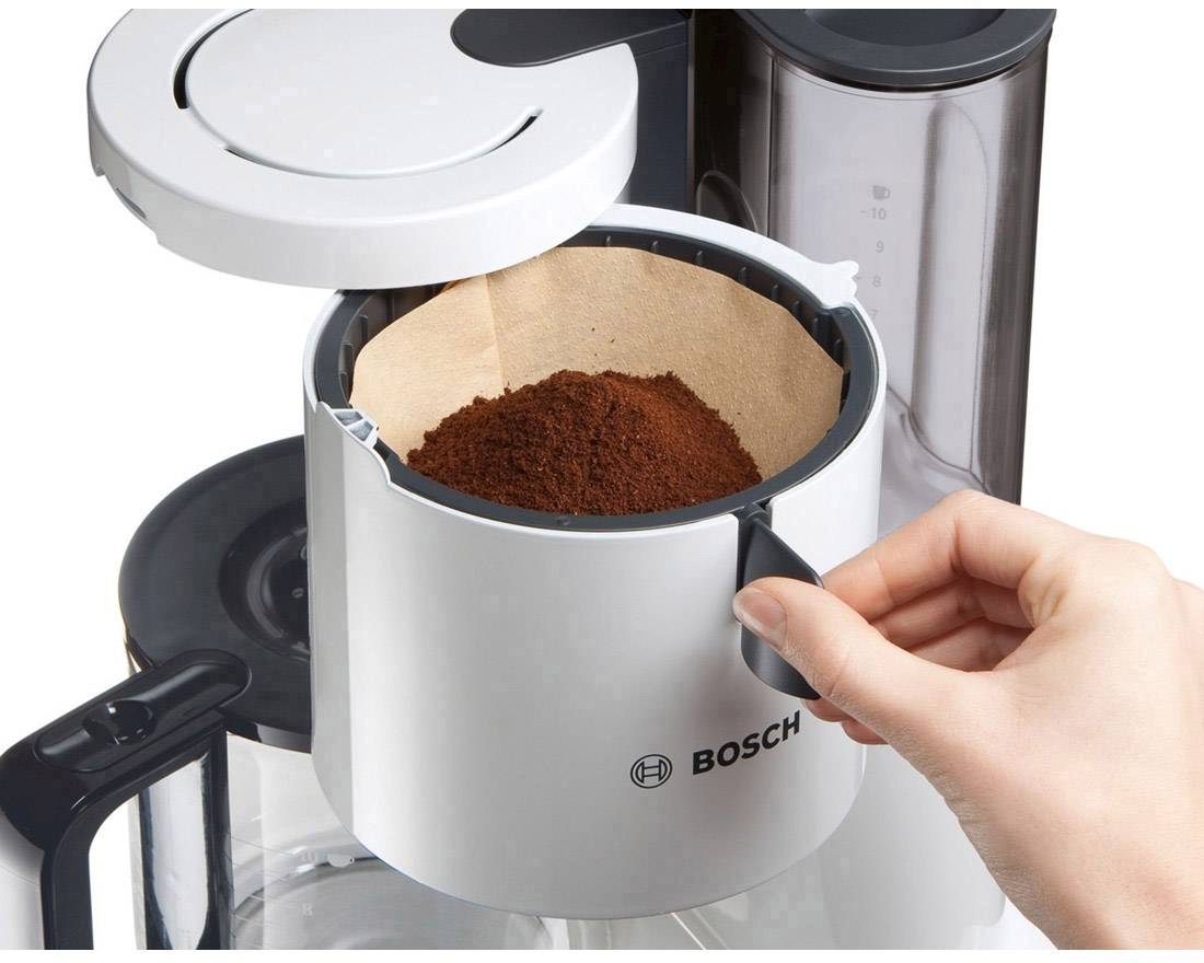 Eine Hand hält den Filterhalter einer Bosch-Kaffeemaschine, gefüllt mit Kaffeepulver, über einer geöffneten Glaskanne.