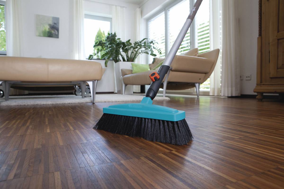 Ein Besen mit blauem Kopf fegt über einen Holzboden in einem Wohnzimmer mit hellen Möbeln und Pflanzen im Hintergrund.