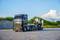 Ein schwarzer Spielzeug-LKW transportiert einen gelben Bagger auf einem Anhänger, vor einem grasbewachsenen Hintergrund unter blauem Himmel.