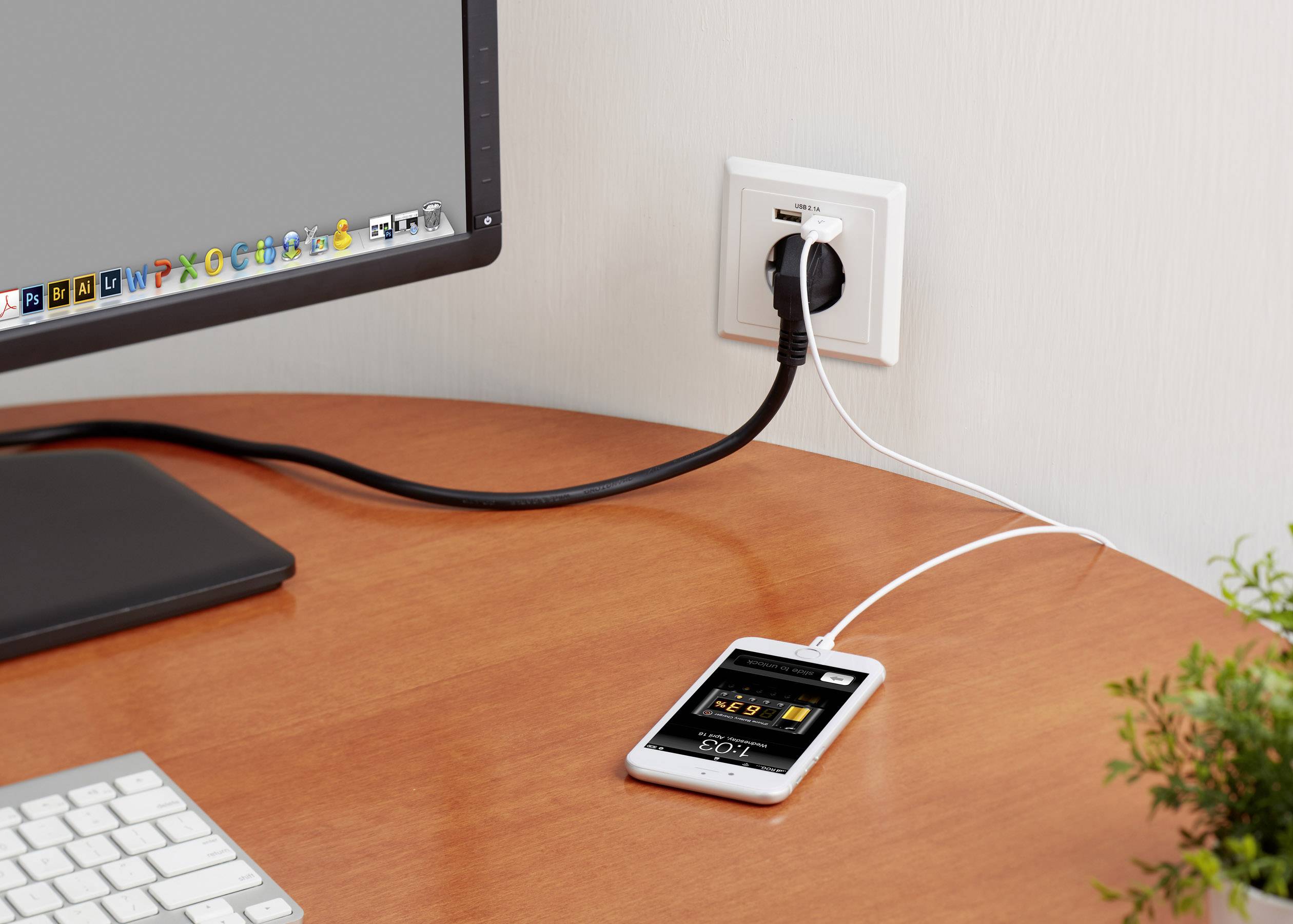 Ein Smartphone wird mit einem Kabel an eine Steckdose angeschlossen und liegt auf einem Tisch neben einem eingeschalteten Computerbildschirm.