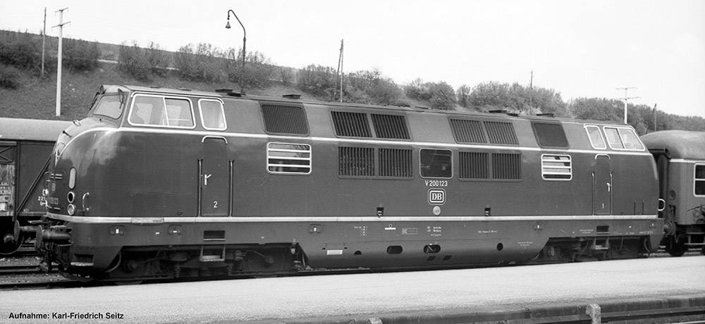 Eine historische Lokomotive der Deutschen Bundesbahn, Modell V 200, steht auf einem Bahngleis. Im Hintergrund sind Bäume und ein Hügel sichtbar.