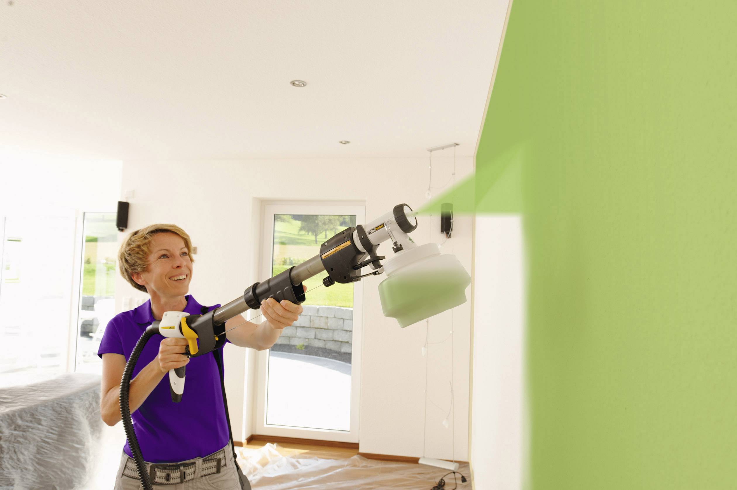 Eine Person sprüht grüne Farbe auf eine Innenwand mit einem Farbsprühsystem in einem hellen Raum.