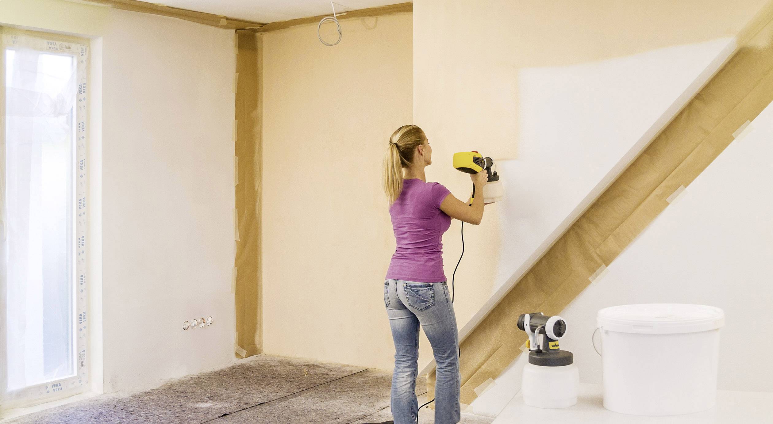 Eine Frau streicht mit einem Farbsprüher eine Wand weiß in einem Raum, der frisch renoviert wird. Werkzeug und Farbeimer stehen daneben.