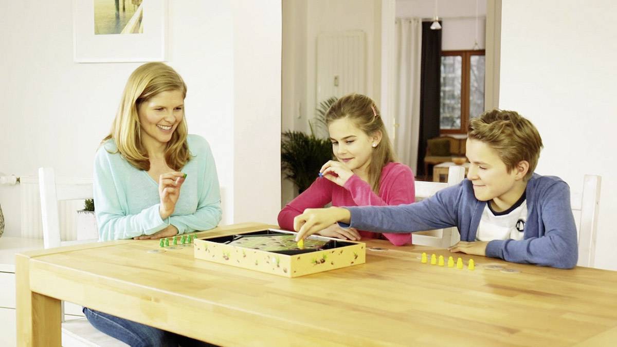 Eine Frau und zwei Kinder spielen ein Brettspiel an einem Holztisch in einem Wohnzimmer. Sie alle lächeln und sind vertieft im Spiel.