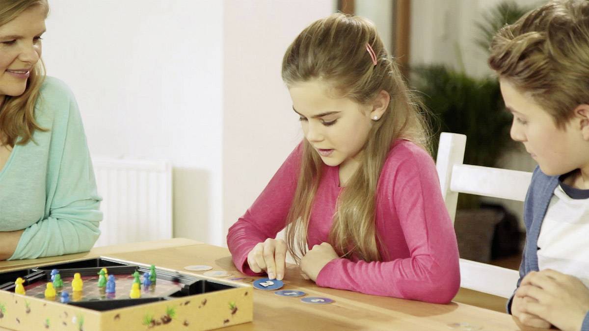 Ein Mädchen spielt ein Brettspiel mit zwei anderen Personen an einem Tisch. Alle wirken konzentriert und engagiert.