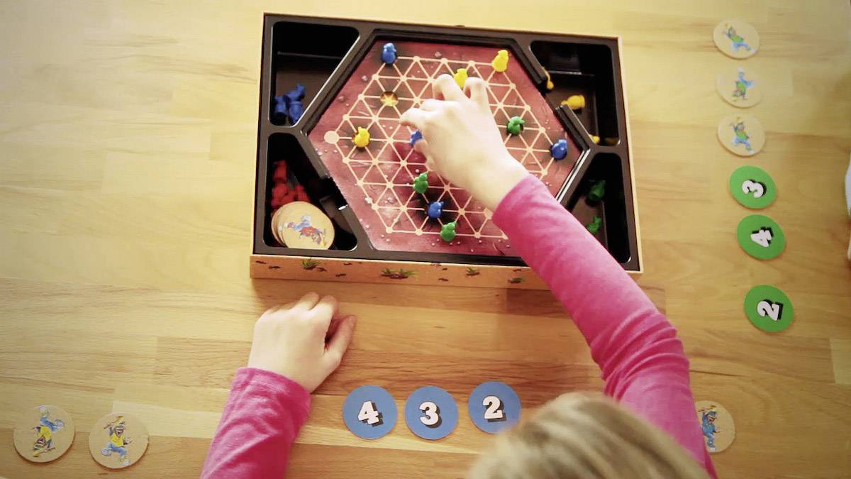 Ein Kind spielt ein Brettspiel, bei dem bunte Spielfiguren auf einem sechseckigen Spielfeld bewegt werden. Zahlenchips liegen daneben.