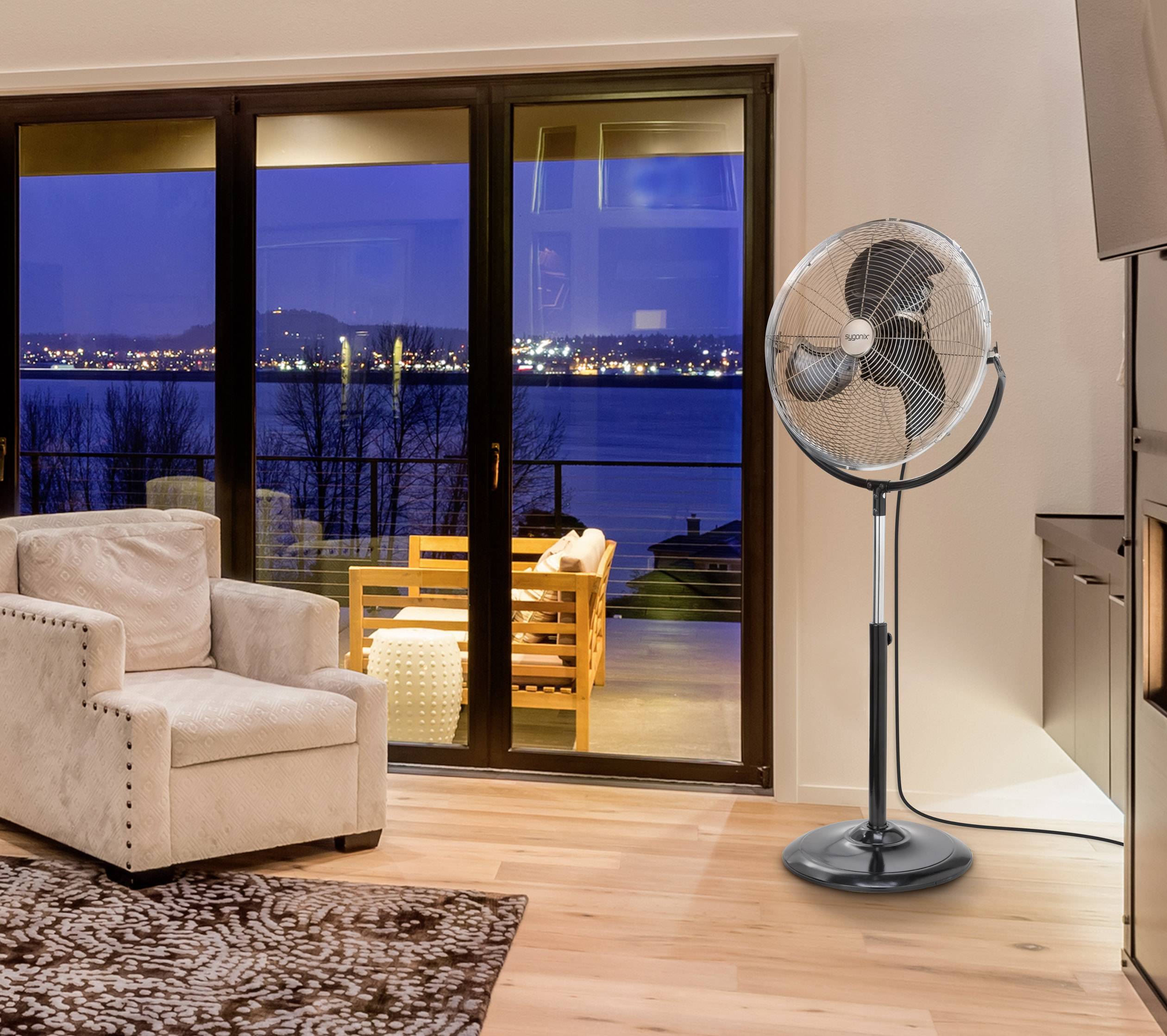 Wohnzimmer mit beigem Sessel, Teppich, und einem Standventilator. Durch die Fensterfront ist ein Balkon mit Aussicht auf einen See bei Nacht sichtbar.
