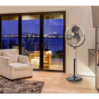 Wohnzimmer mit beigem Sessel, Teppich, und einem Standventilator. Durch die Fensterfront ist ein Balkon mit Aussicht auf einen See bei Nacht sichtbar.