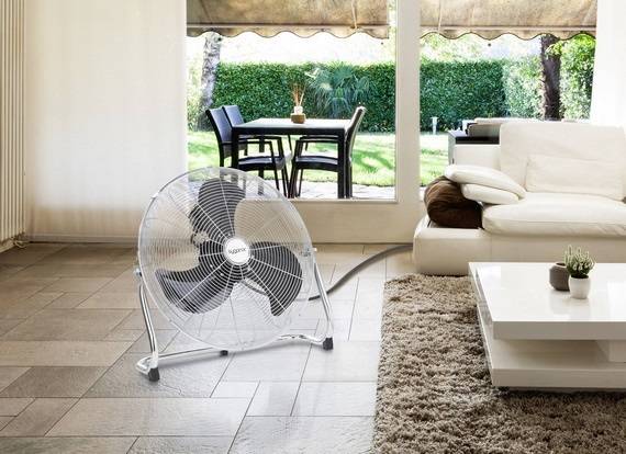 Ein Ventilator steht auf einem Fliesenboden in einem Wohnzimmer. Durch eine Fensterfront sieht man einen Garten mit Tisch und Stühlen.