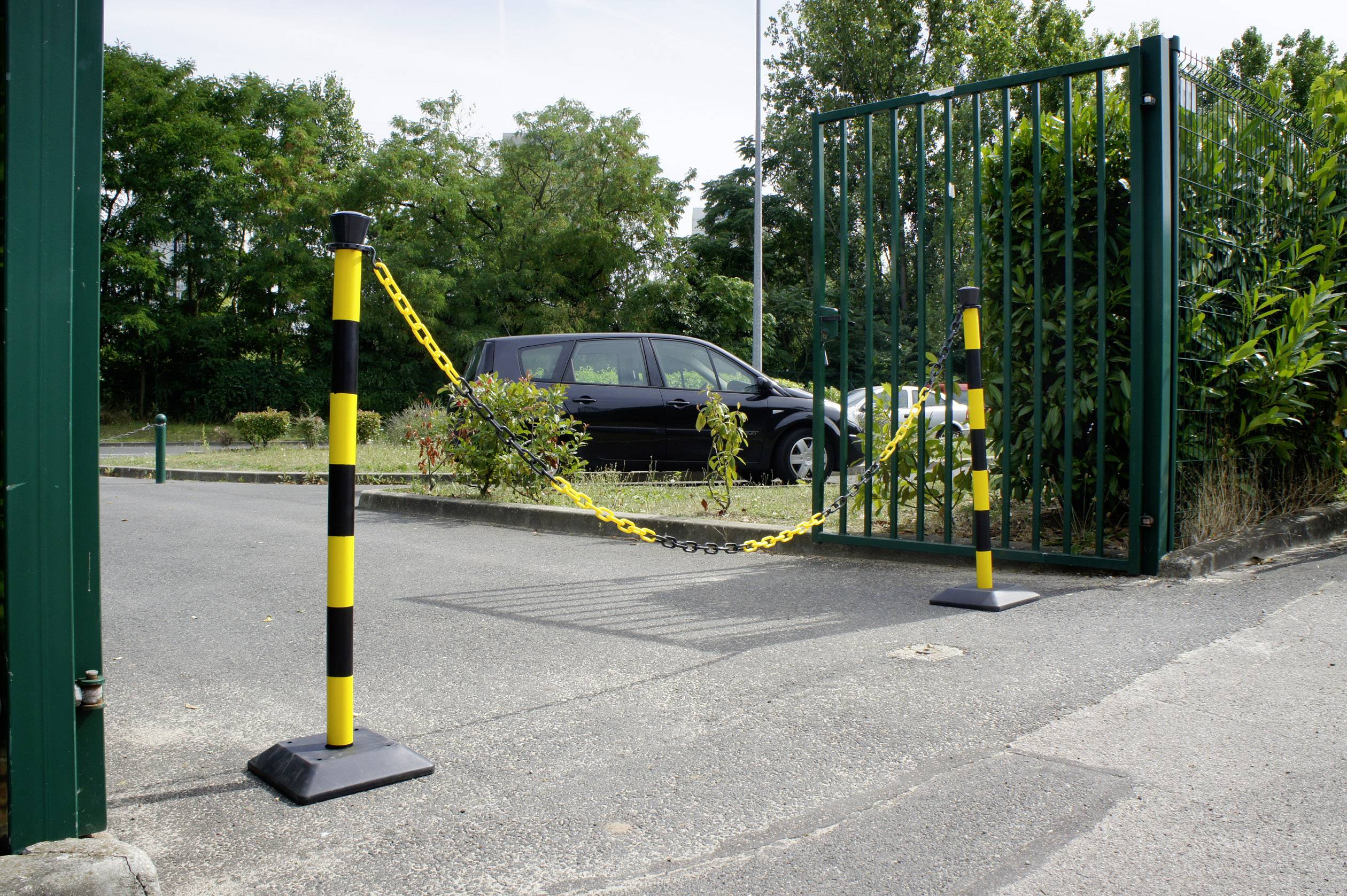 Ein offenes Tor mit einer gelb-schwarzen Sicherheitskette über einem Zufahrtsweg, dahinter ein geparktes Auto und grüne Büsche.