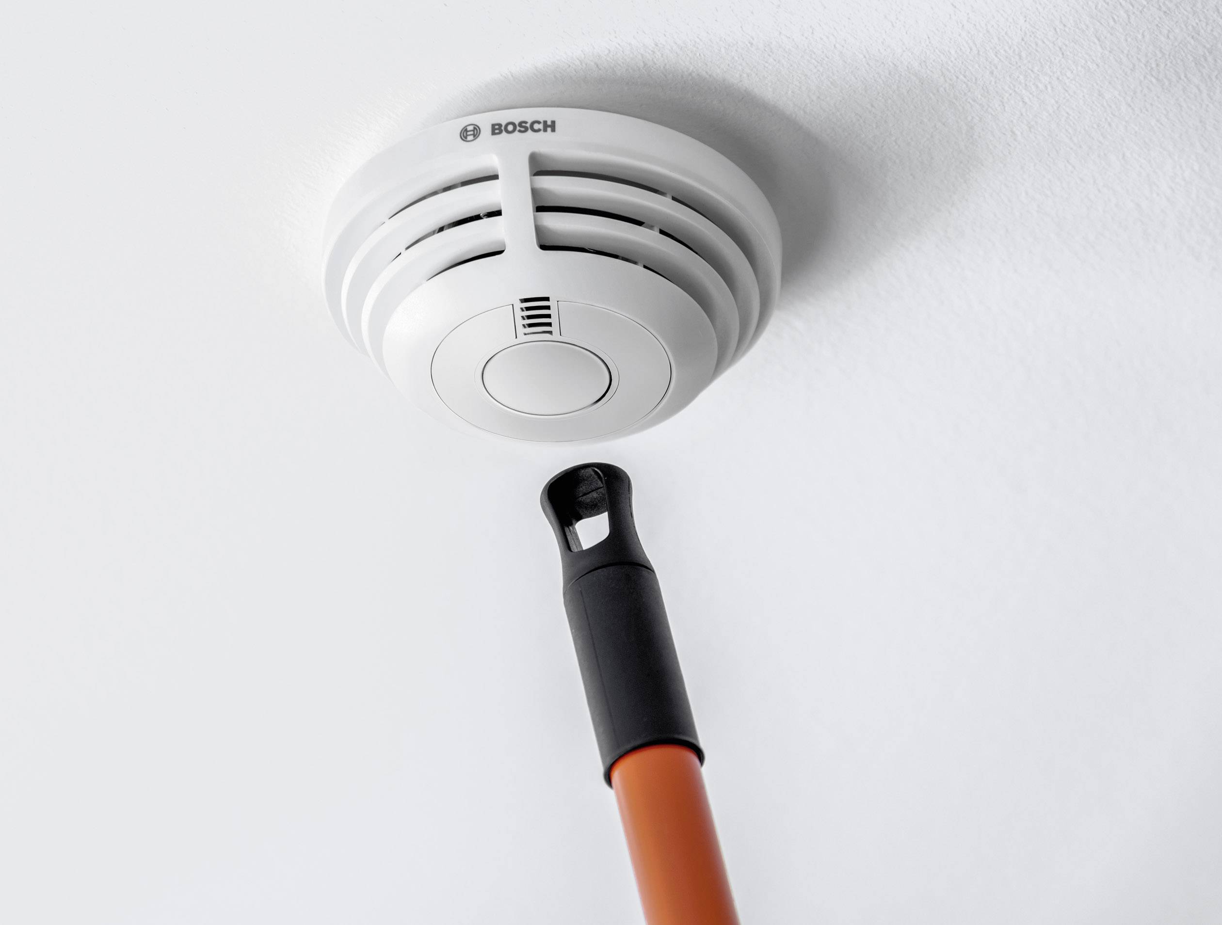 Ein Rauchmelder an der Decke wird mit einem Stab getestet. Er ist weiß und rund, die Marke ist sichtbar.