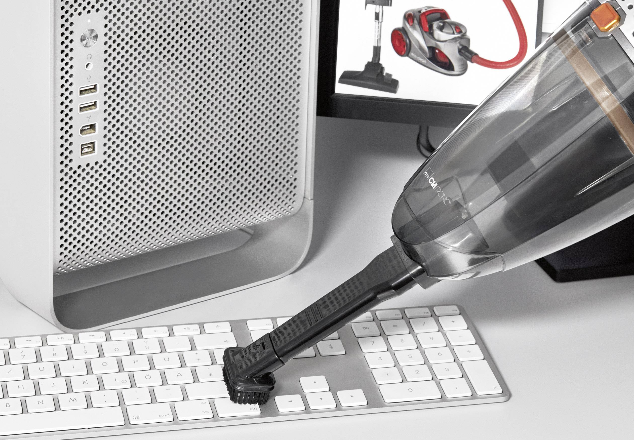 Ein Handstaubsauger reinigt die Tastatur eines Computers. Im Hintergrund sind ein Computergehäuse und zwei Bildschirme sichtbar.