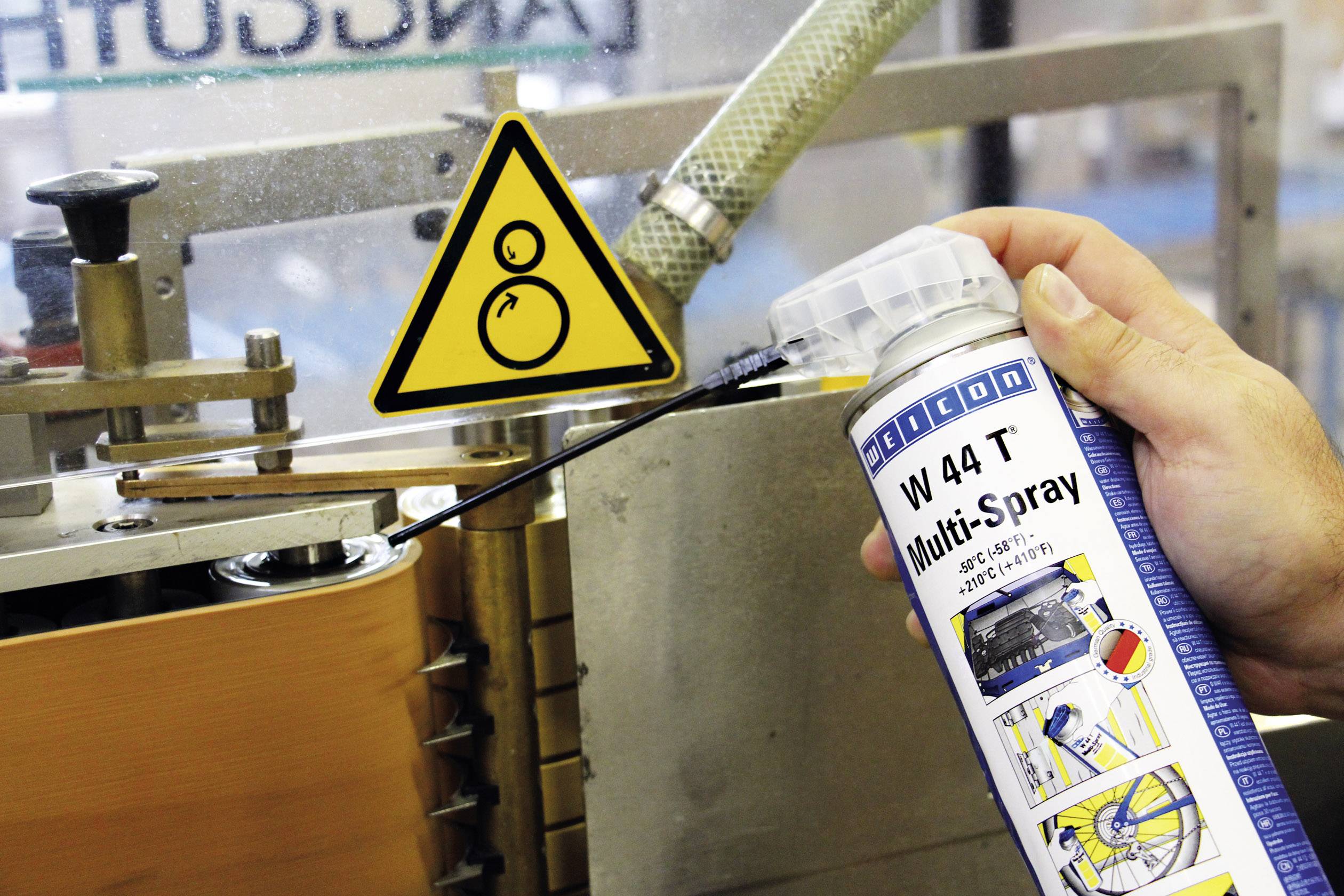 Eine Person hält eine Spraydose mit der Aufschrift 'W 44 T Multi-Spray' vor einer Maschine. Ein Warnschild zeigt 'Achtung'.