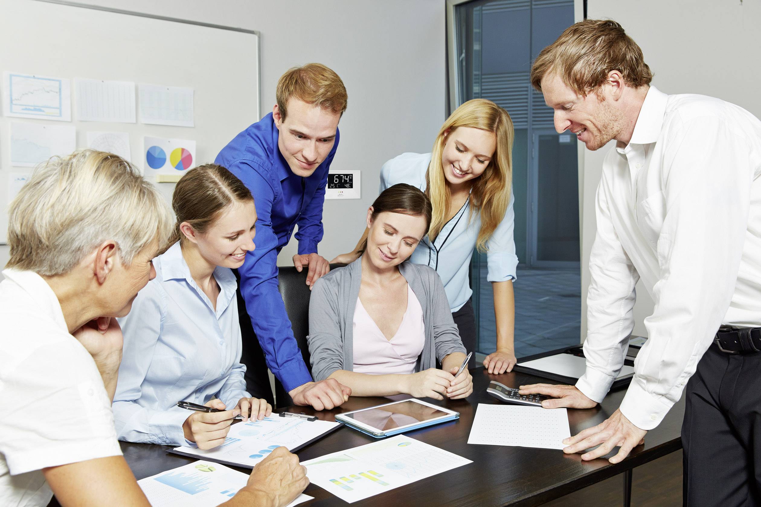 Sechs Personen in einem Büro besprechen Dokumente und Diagramme. Eine Person zeigt auf ein Tablet, während andere aufmerksam zuhören.