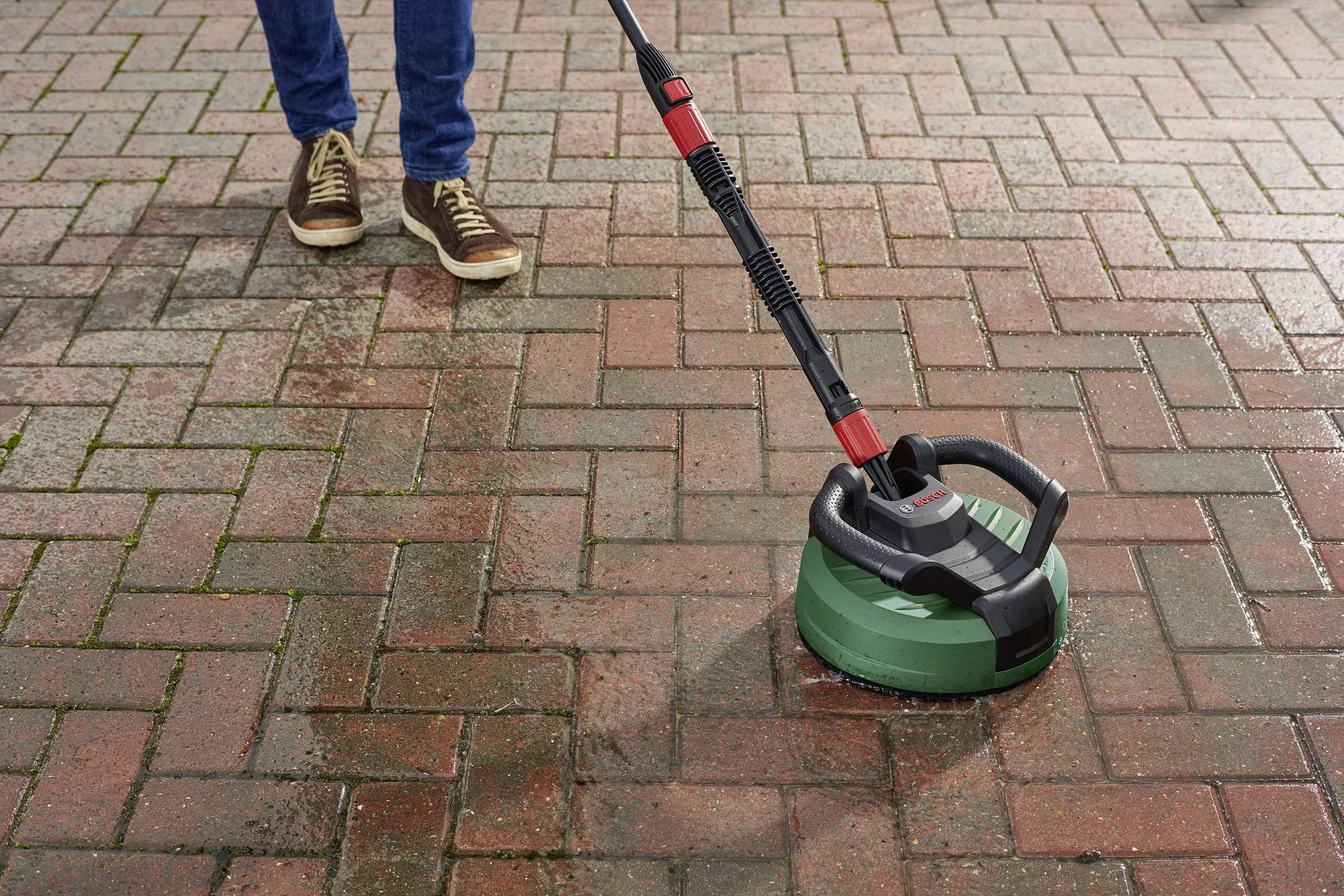 Eine Person reinigt mit einem Hochdruckreiniger einen Steinboden. Links ist der gereinigte Boden heller, rechts noch dunkel.