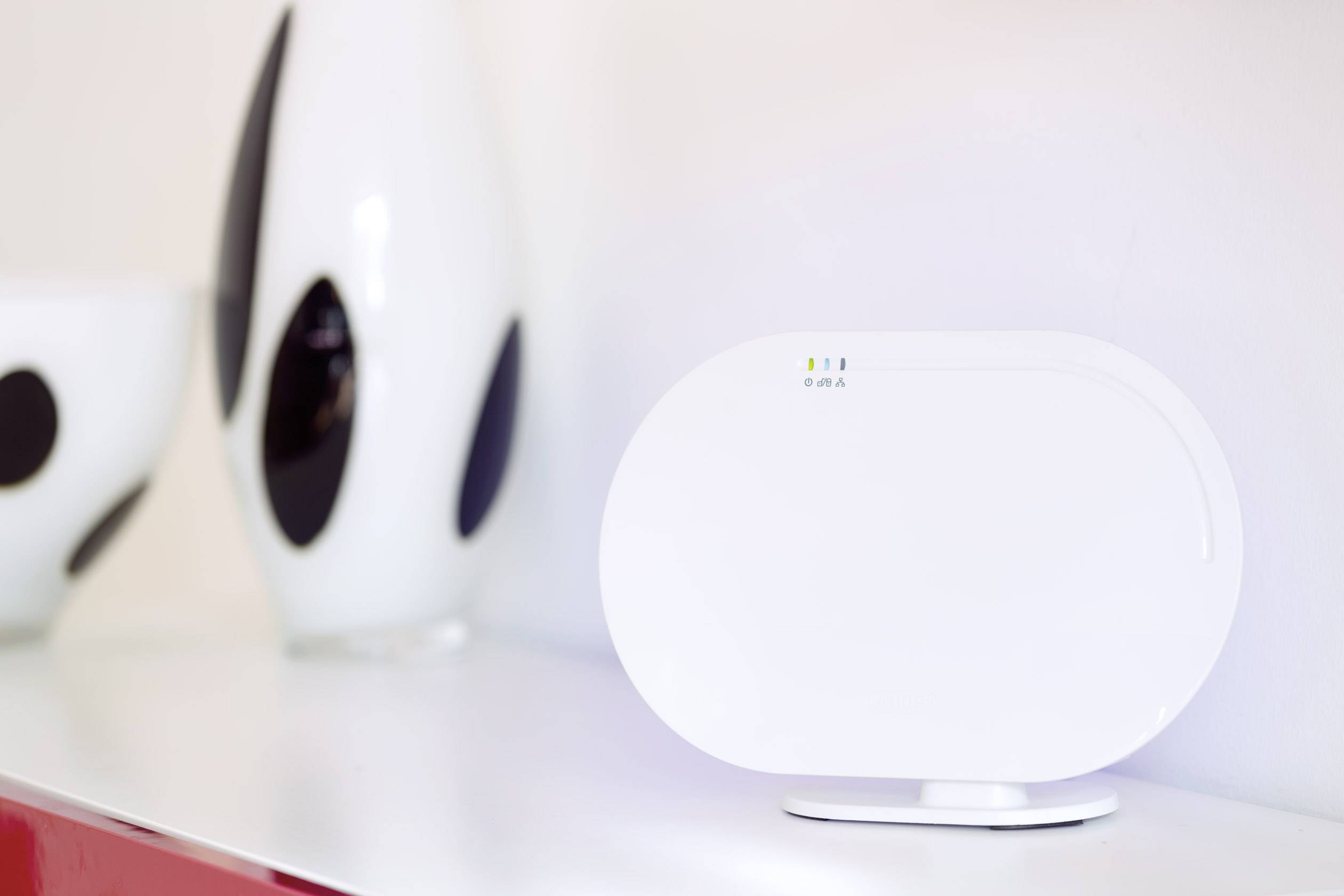Ein weißes, ovales Wi-Fi-Gerät steht auf einem Schreibtisch. Im Hintergrund sind dekorative Vasen zu sehen.