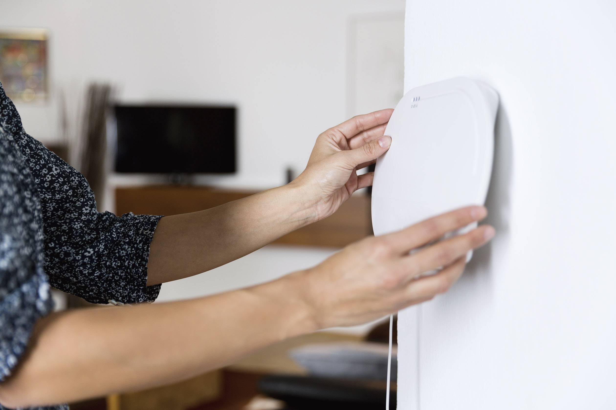 Eine Person montiert ein quadratisches Gerät an einer weißen Wand in einem Wohnzimmer. Ein Fernseher ist im Hintergrund sichtbar.