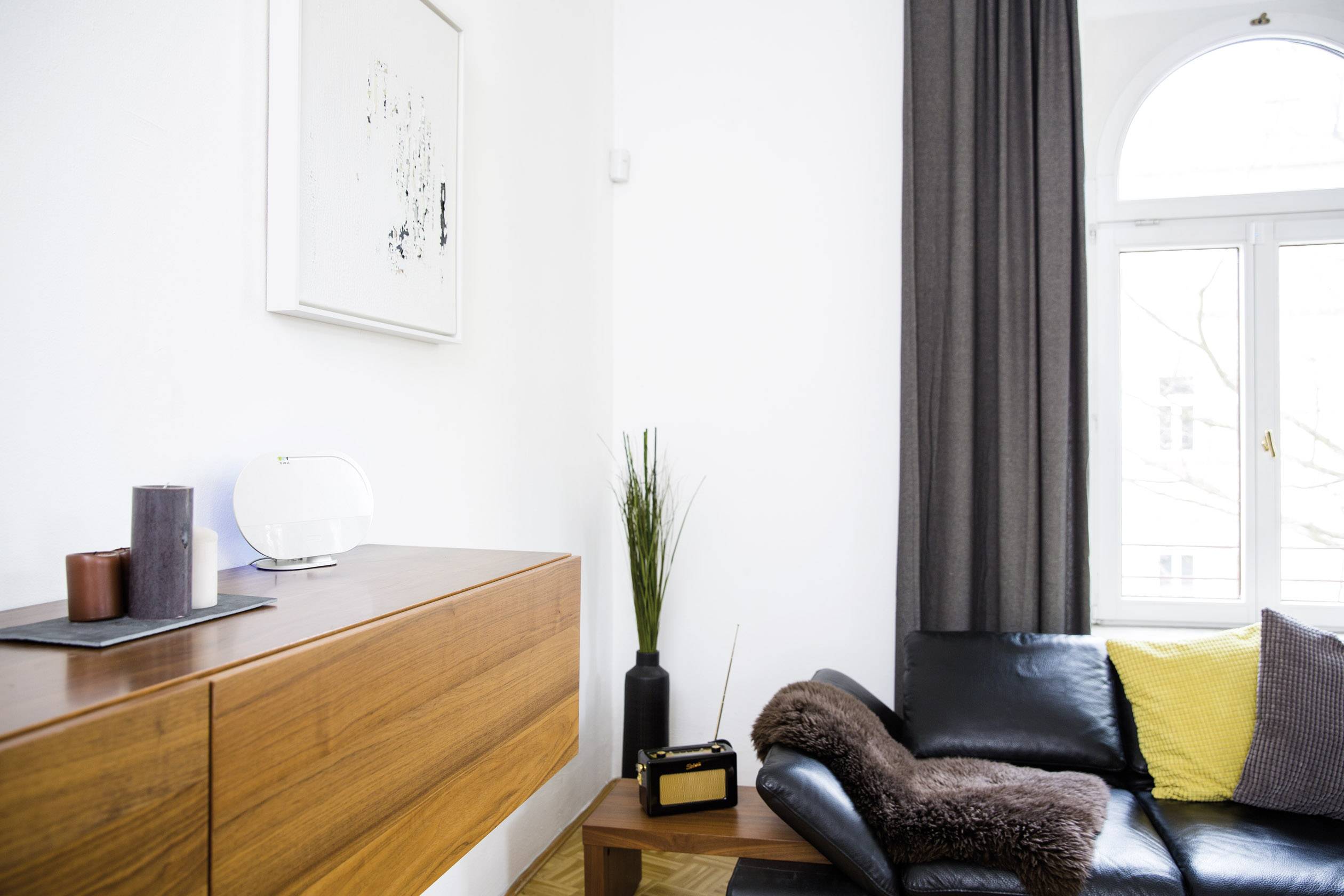 Wohnzimmer mit schwarzem Ledersofa, gelbem Kissen, Holzschrank und Dekorationen. Helle Wände und großes Fenster rechts.