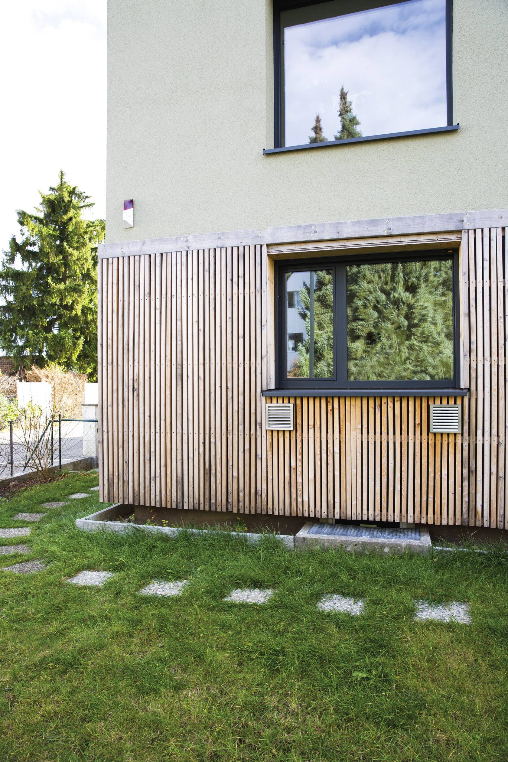 Ein modernes Gebäude mit Holzelementen, großem Fenster und grüner Wiese, umgeben von Bäumen. Entspanntes, naturnahes Ambiente.