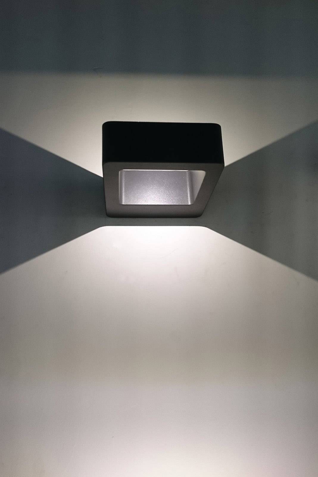 Eine moderne Wandlampe wirft symmetrische Licht- und Schattenstrahlen auf eine graue Wand.