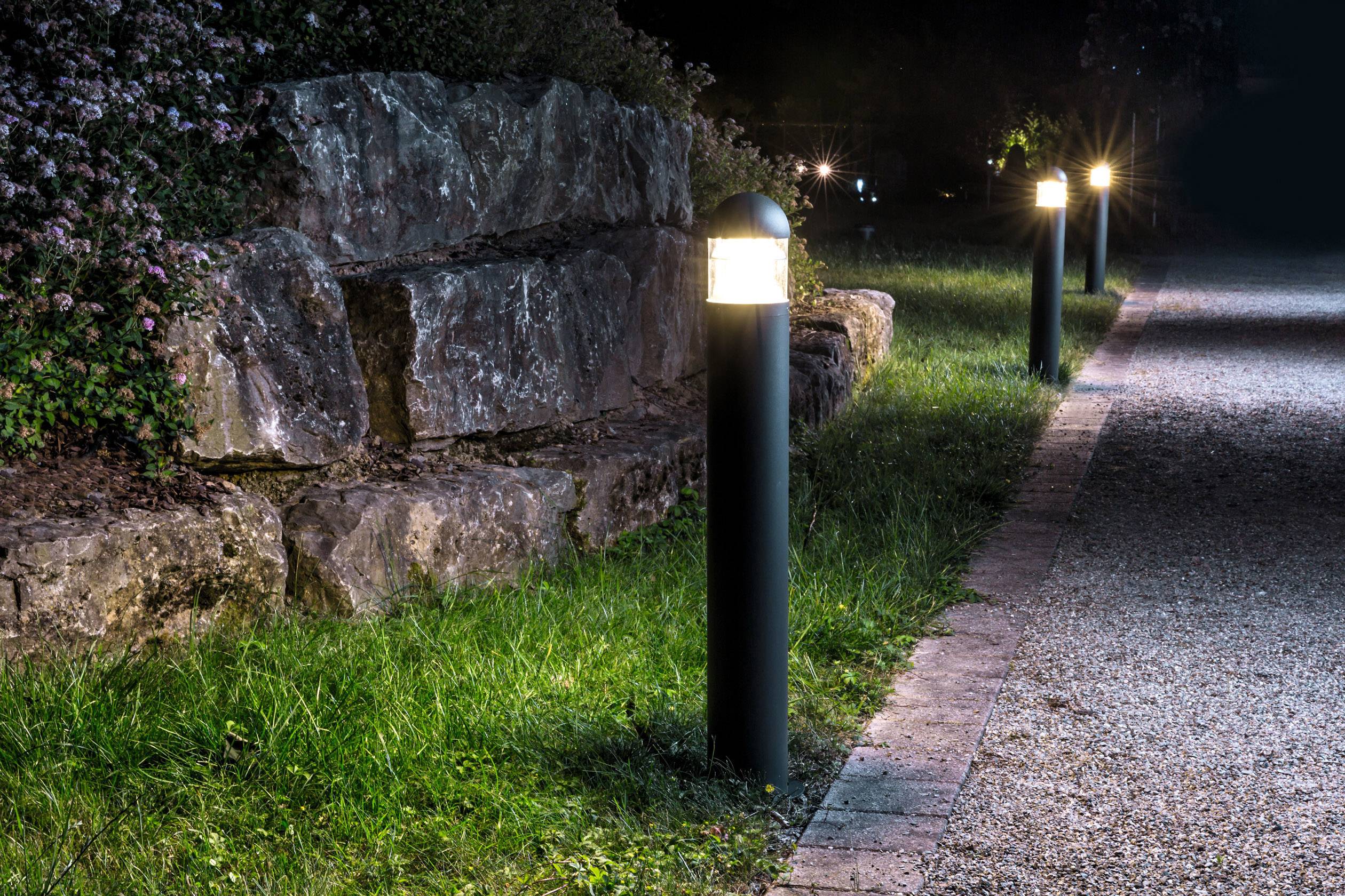 Ein nächtlicher Gartenweg mit leuchtenden Wegleuchten, die den Weg entlang eines gepflasterten Pfads neben einer Steinmauer beleuchten.