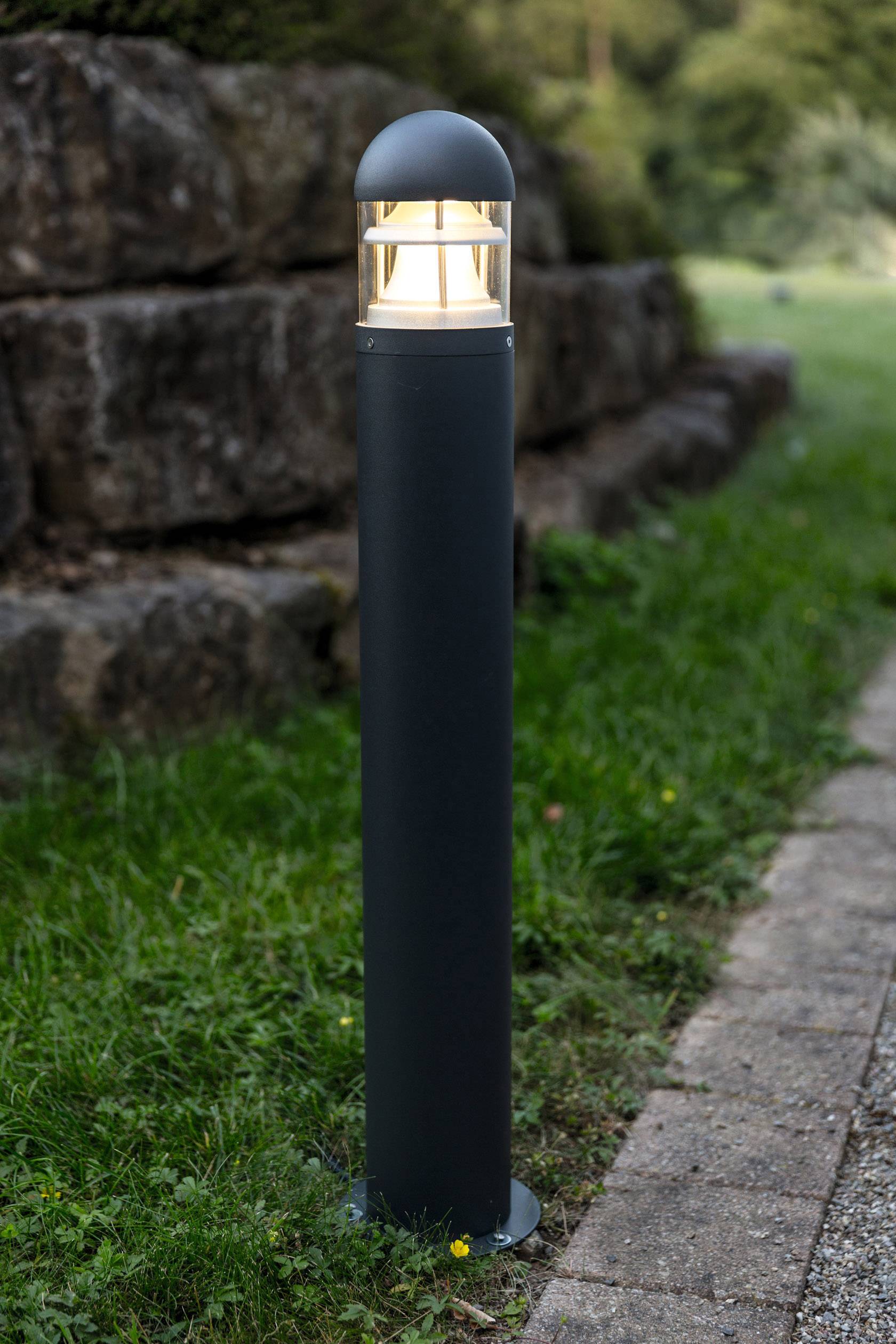 Eine schwarze Gartenlampe leuchtet neben einem gepflasterten Weg, umgeben von Gras und Steinmauern in einem Außenbereich.