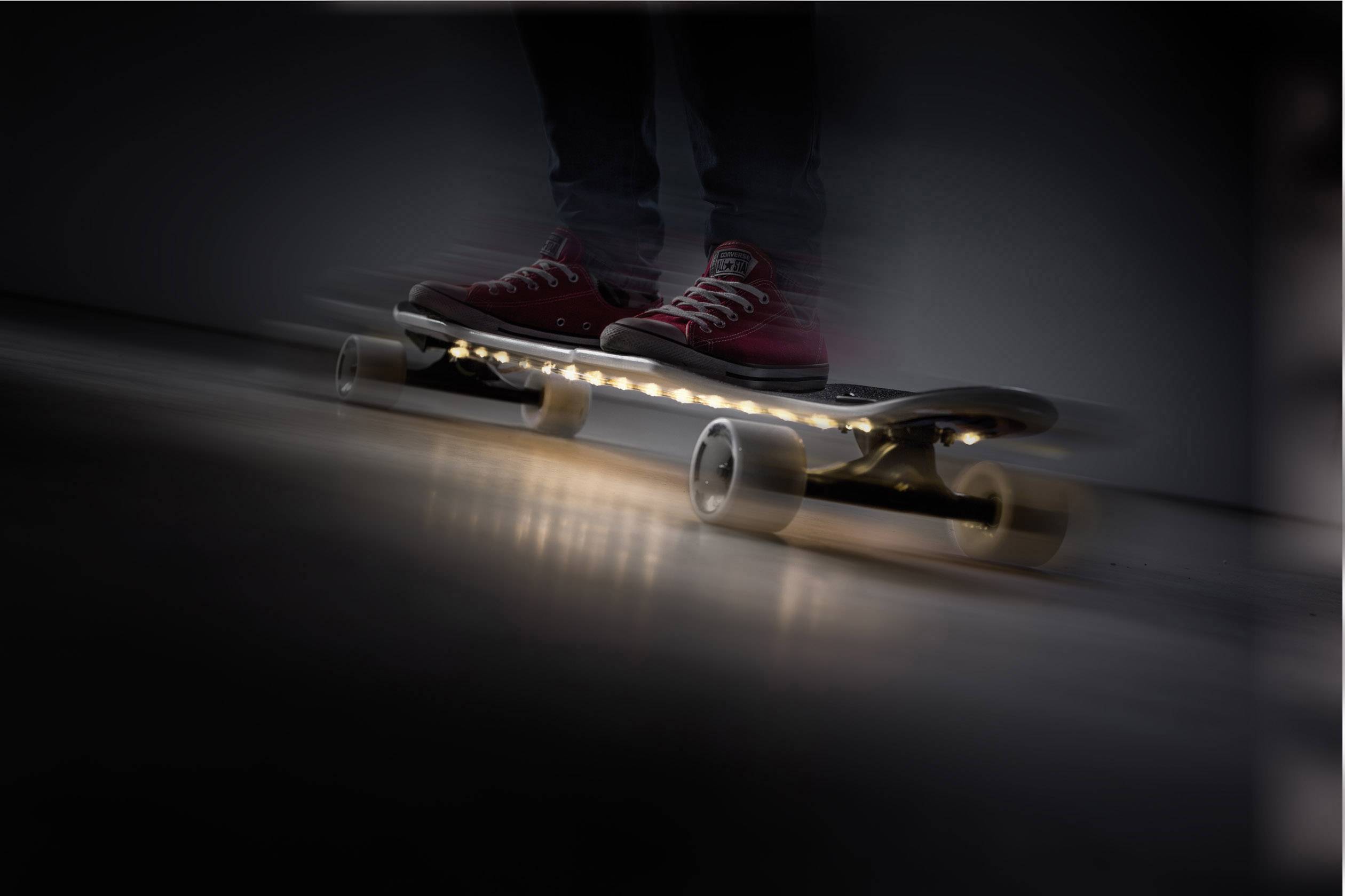 Jemand steht auf einem beleuchteten Skateboard, dessen Lichter bei Bewegung verschwommen wirken. Die Person trägt rote Turnschuhe.