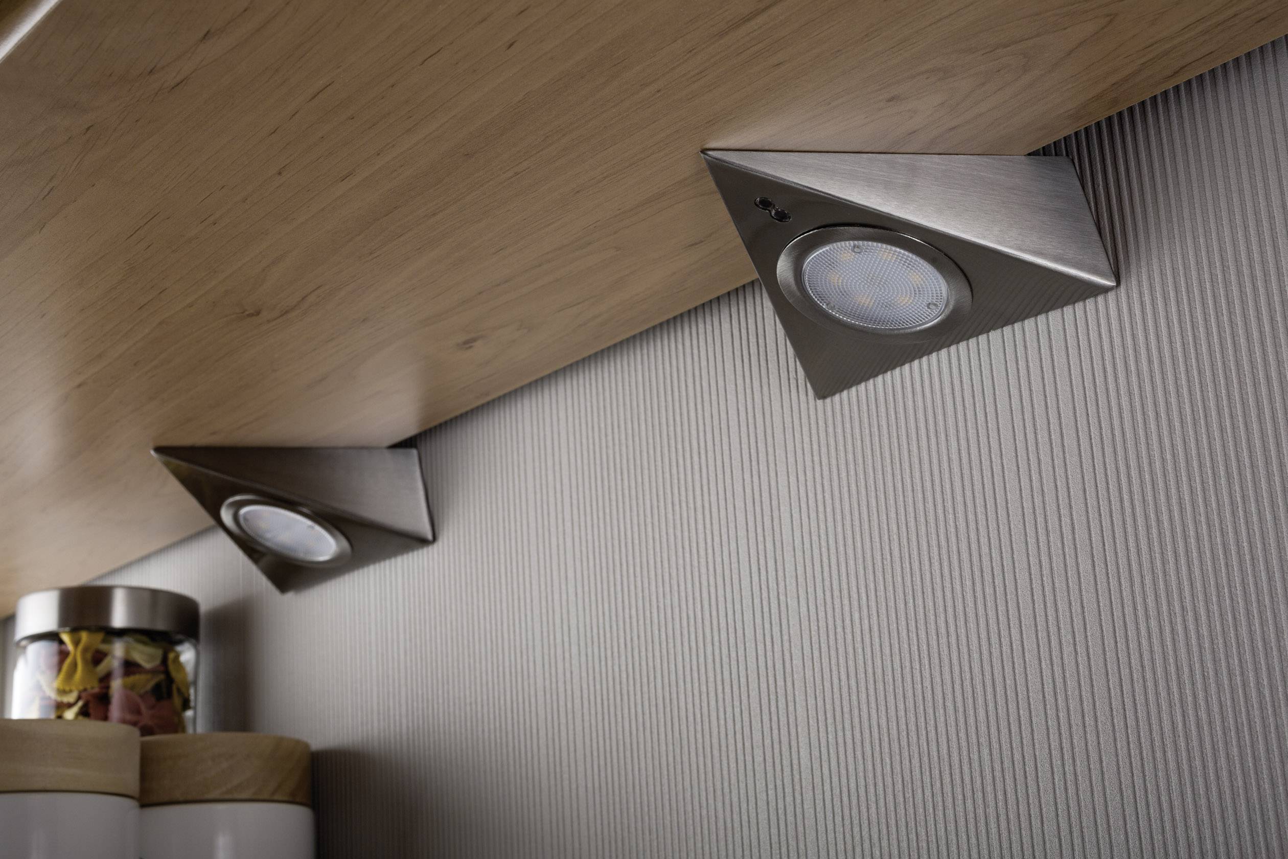 Zwei dreieckige LED-Unterbauleuchten aus Metall sind unter einem Holzregal montiert und beleuchten den Bereich darunter.