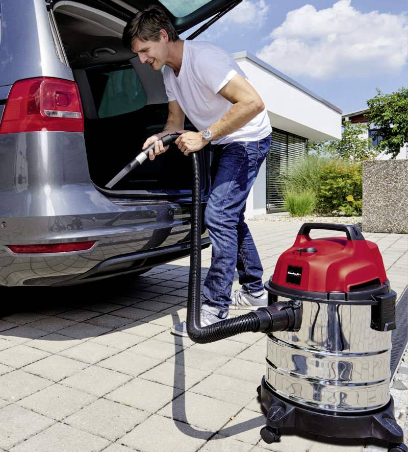Ein Mann reinigt den Kofferraum seines Autos mit einem Nass-Trockensauger auf einer Einfahrt vor einem modernen Haus bei sonnigem Wetter.