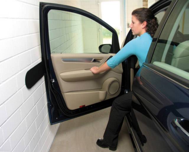 Eine Frau in blauer Bluse öffnet vorsichtig die Tür eines Autos, das dicht neben einer weißen Ziegelwand geparkt ist.