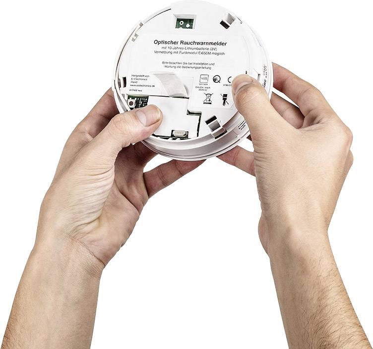 Zwei Hände halten einen optischen Rauchwarnmelder. Das Gerät ist geöffnet, und auf der Rückseite sind ein Batteriefach sowie Schalter sichtbar.