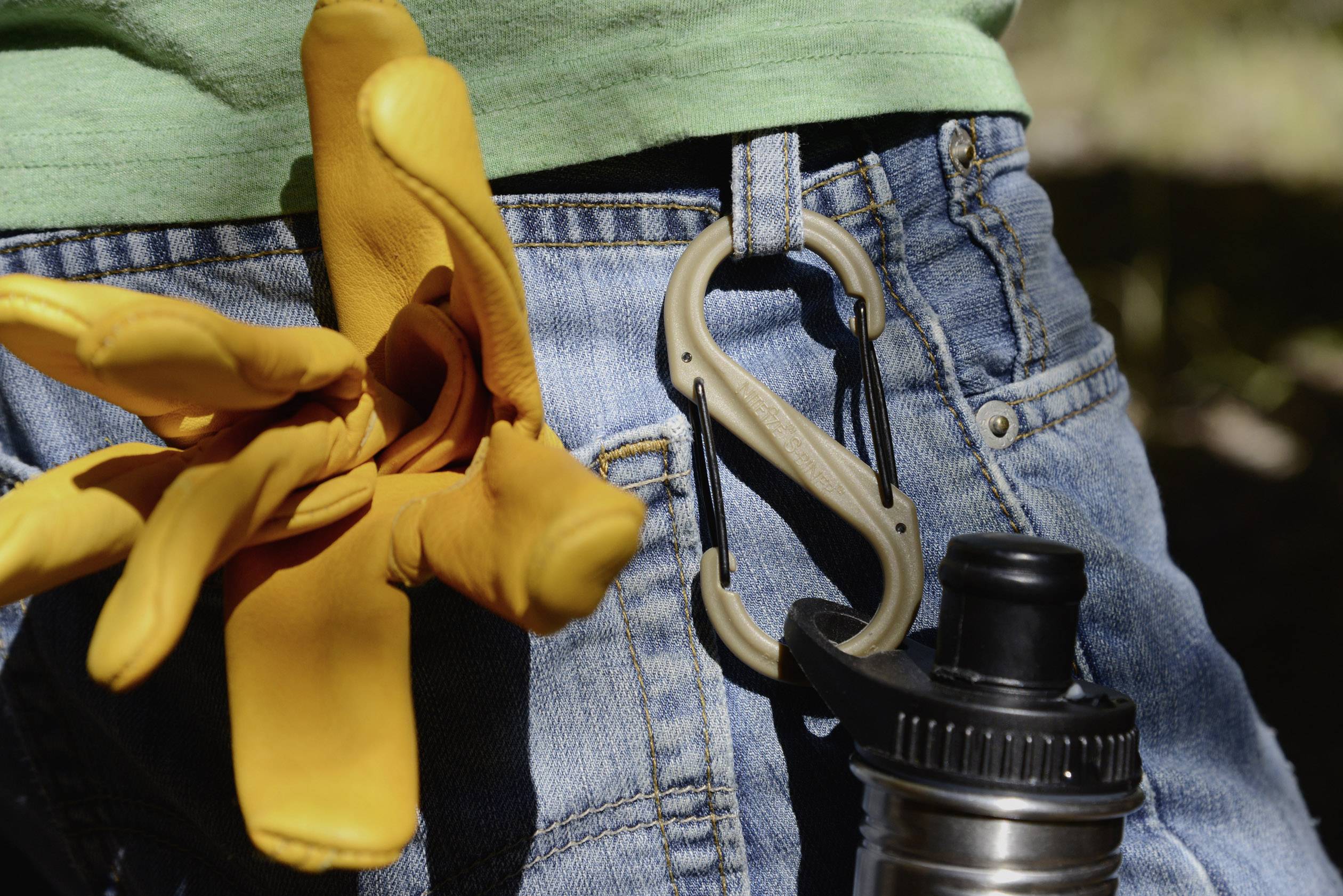 Gelbe Arbeitshandschuhe in einer Jeanshintertasche, neben einem Karabinerhaken und einer Trinkflasche aus Metall. Ideal für Outdoor-Aktivitäten.