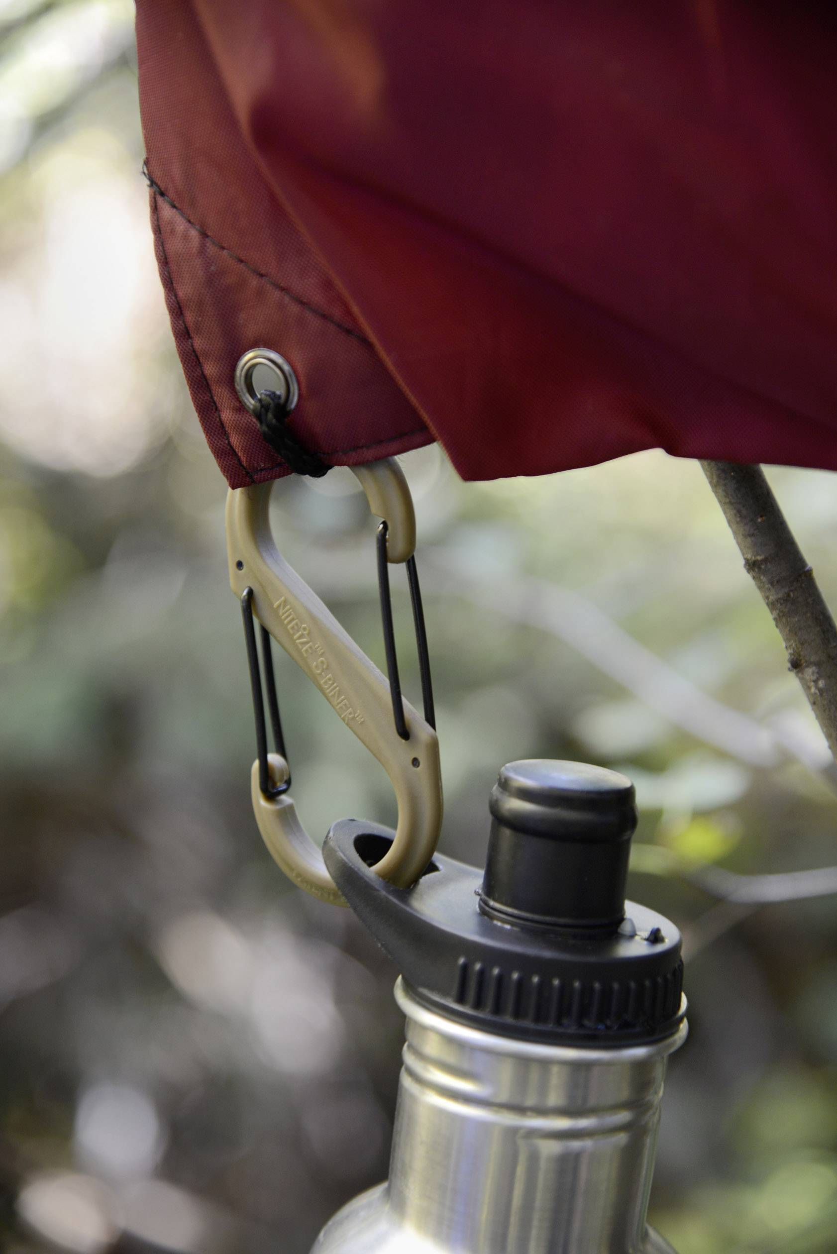 Eine rote Plane wird mit einem Karabinerhaken an einem Ast befestigt; darunter hängt eine Edelstahlflasche mit schwarzem Verschluss.