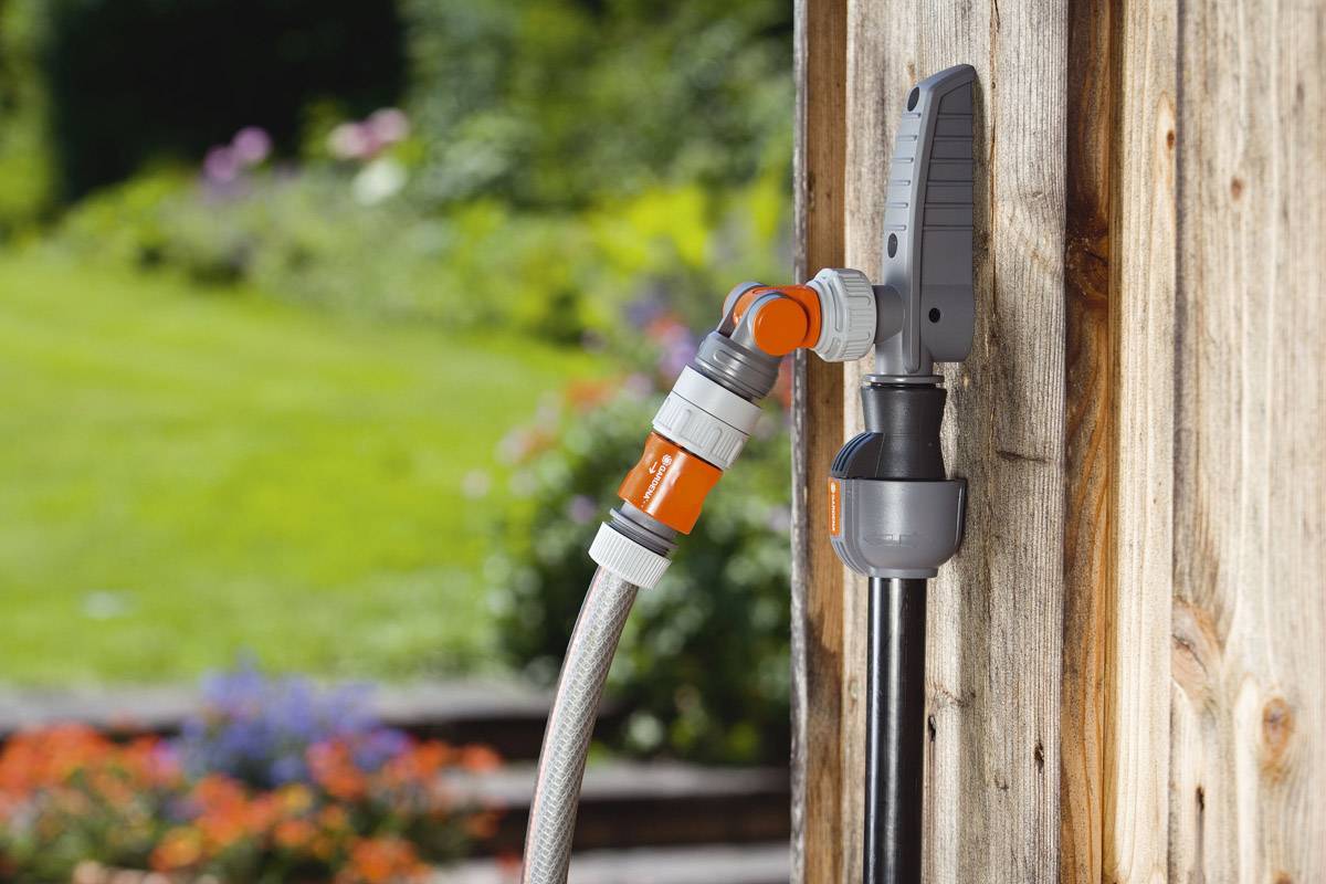 Ein Gartenschlauch mit grauen und orangefarbenen Anschlüssen ist an einen Wasserhahn an einer Holzwand angeschlossen, im Hintergrund ein Garten.