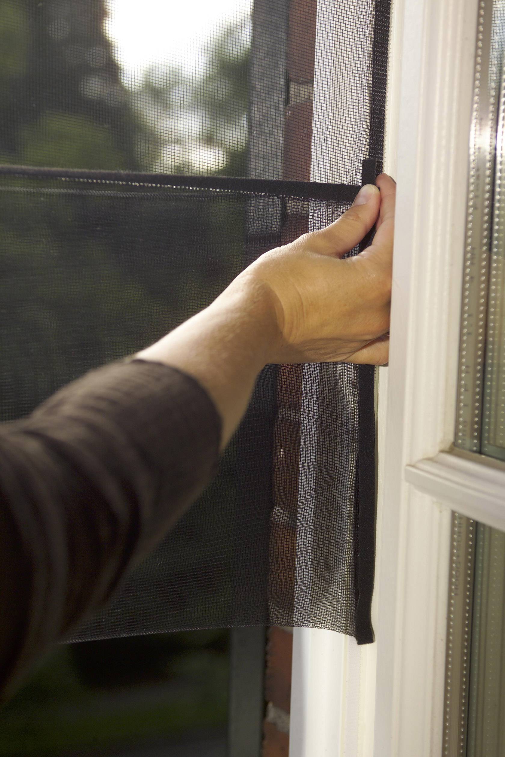 Person legt ein schwarzes Fliegengitter an ein offenes Fenster an, um Insekten draußen zu halten. Fensterszenerie im Hintergrund.