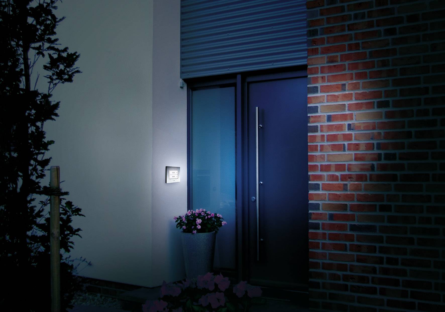 Eingangsbereich eines Hauses bei Nacht, beleuchtet durch Außenlampe neben der Haustür. Daneben steht ein Blumenkübel mit blühenden Pflanzen.
