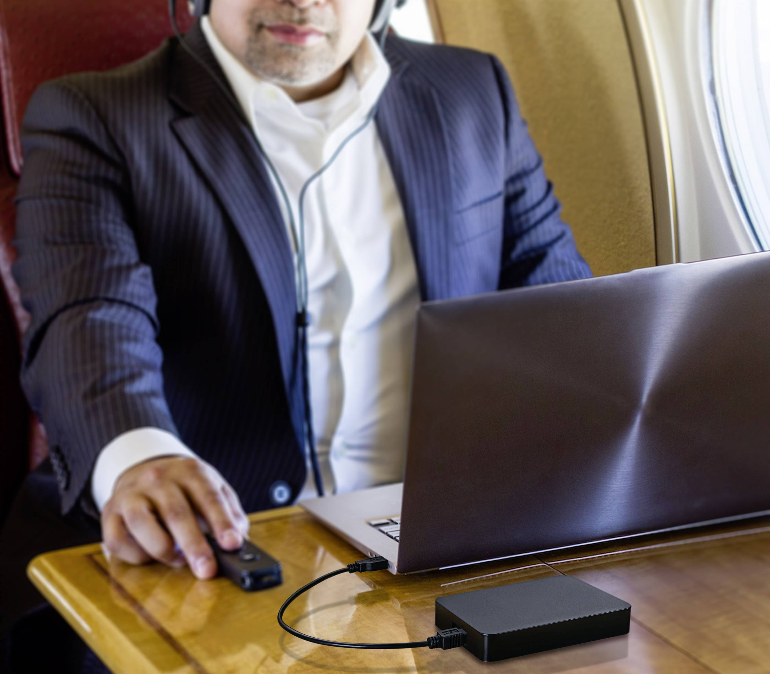 Ein Mann im Anzug arbeitet an einem Laptop in einem Flugzeug, mit einem externen Laufwerk angeschlossen. Er trägt Kopfhörer.