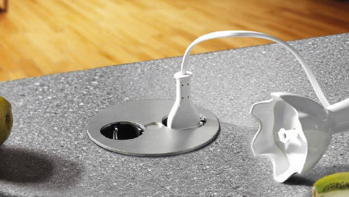 Eine Küchenarbeitsplatte mit eingebauter versenkbarer Steckdose. Ein weißes Kabel ist eingesteckt, ein Mixer-Aufsatz liegt daneben.