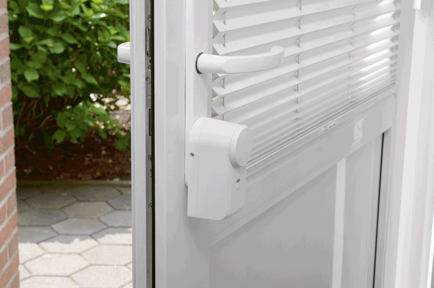 Die Nahaufnahme zeigt eine geöffnete Tür mit integrierter Jalousie und weißem Türgriff. Draußen sind Pflastersteine und Büsche sichtbar.