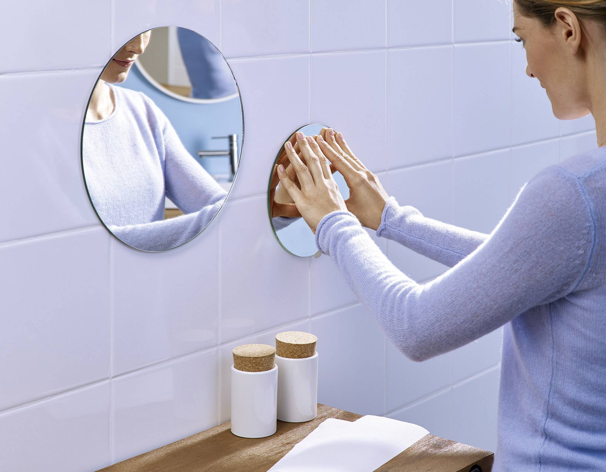 Eine Person wäscht ihre Hände in einem Badezimmer, Spiegelreflexion sichtbar. Auf dem Tisch stehen zwei Behälter.