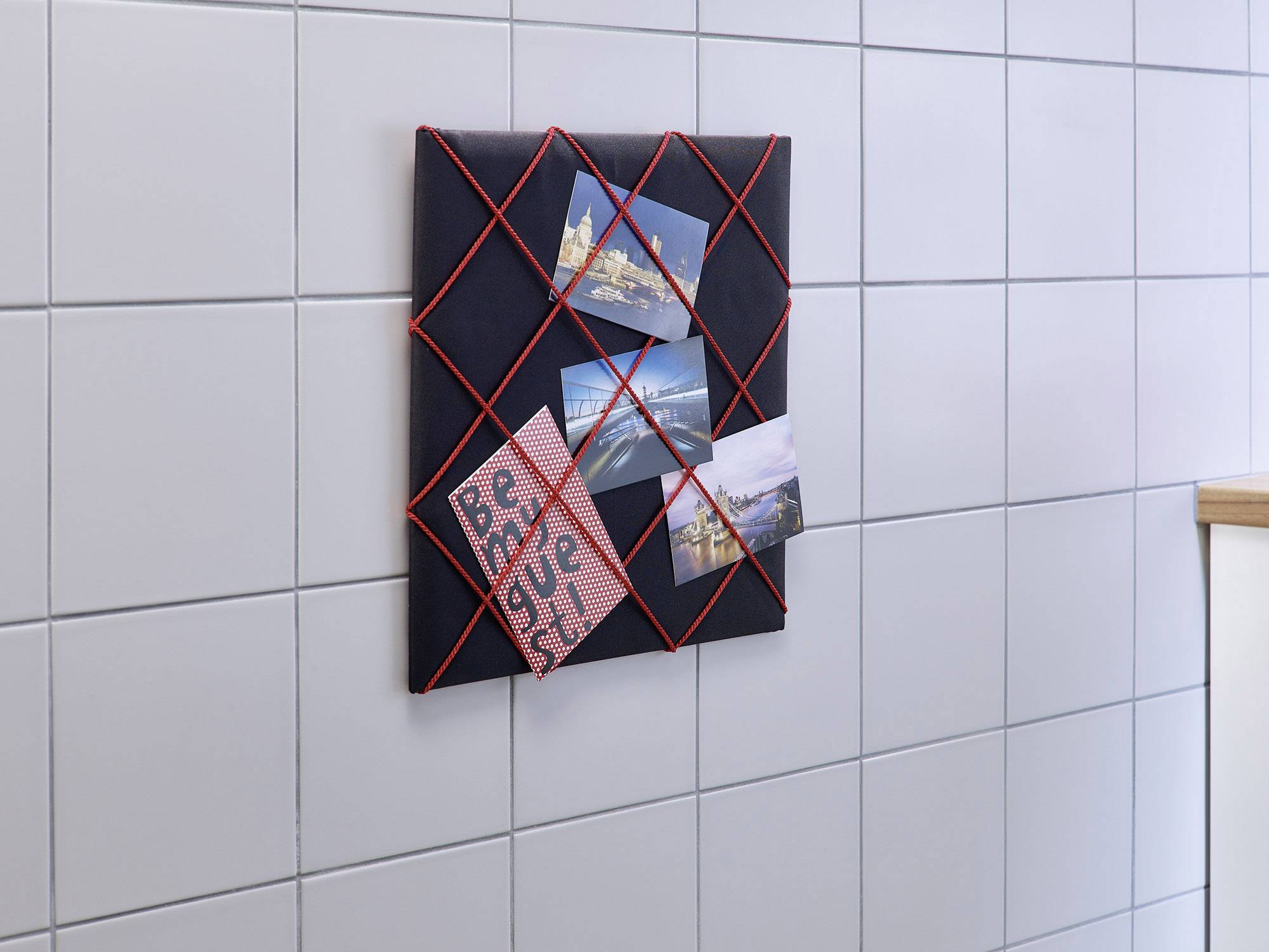 Eine Pinnwand mit roten Fäden an einer weißen gefliesten Wand zeigt eine Postkarte mit 'Be my guest!' und drei Landschaftsfotos.